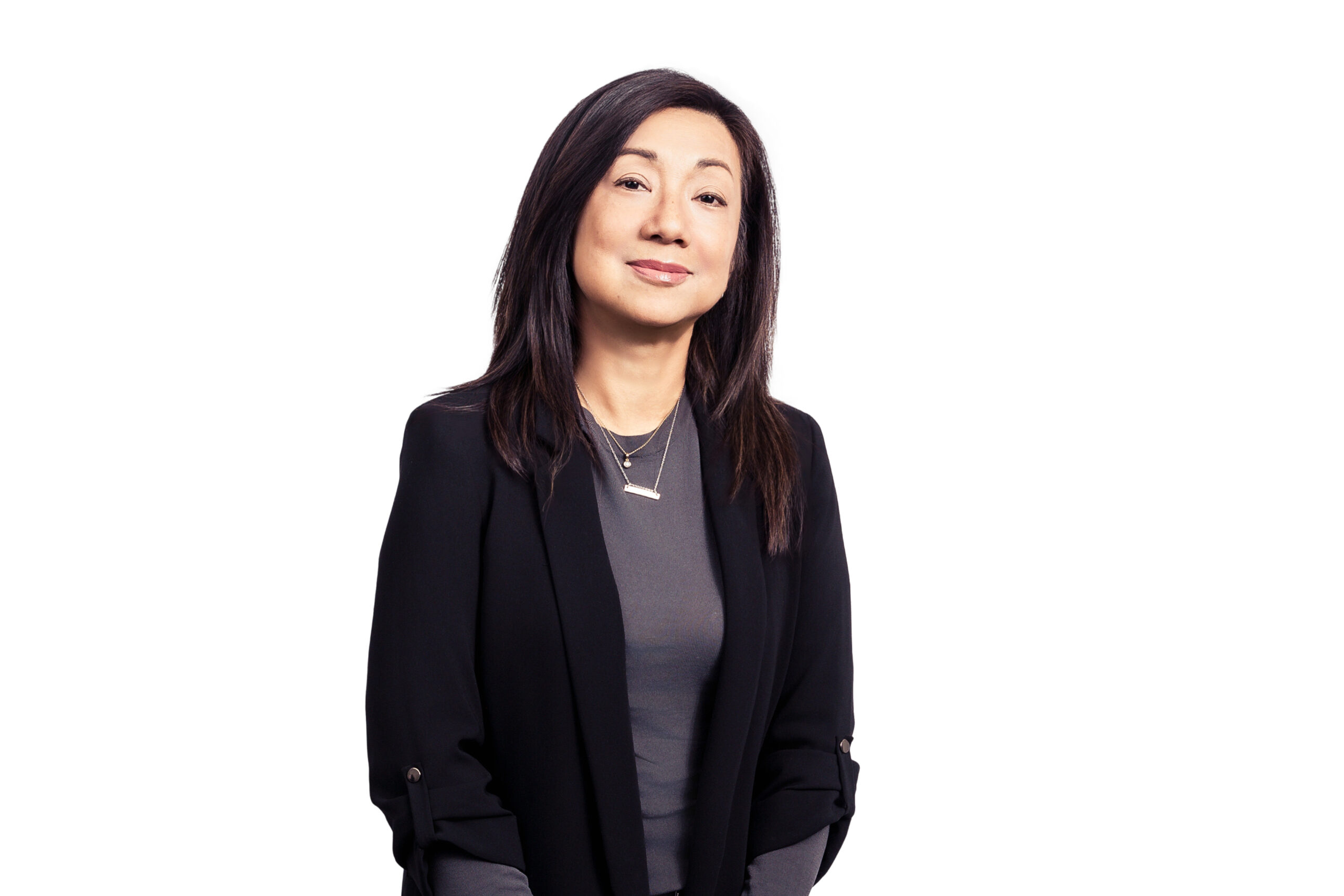 Woman with long dark hair, wearing a black blazer over a gray top, smiling against a white background.
