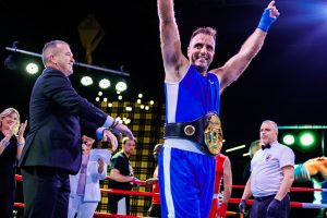 A victorious boxer in blue raises his arms, holding a championship belt, while a judge applauds.