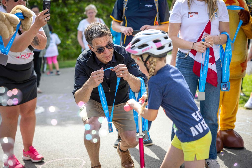 A man presents a medal to a young girl in a helmet, surrounded by cheering participants.