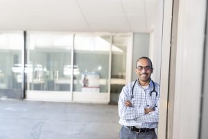 Smiling male doctor in a checked shirt with a stethoscope, standing outside a glass building.