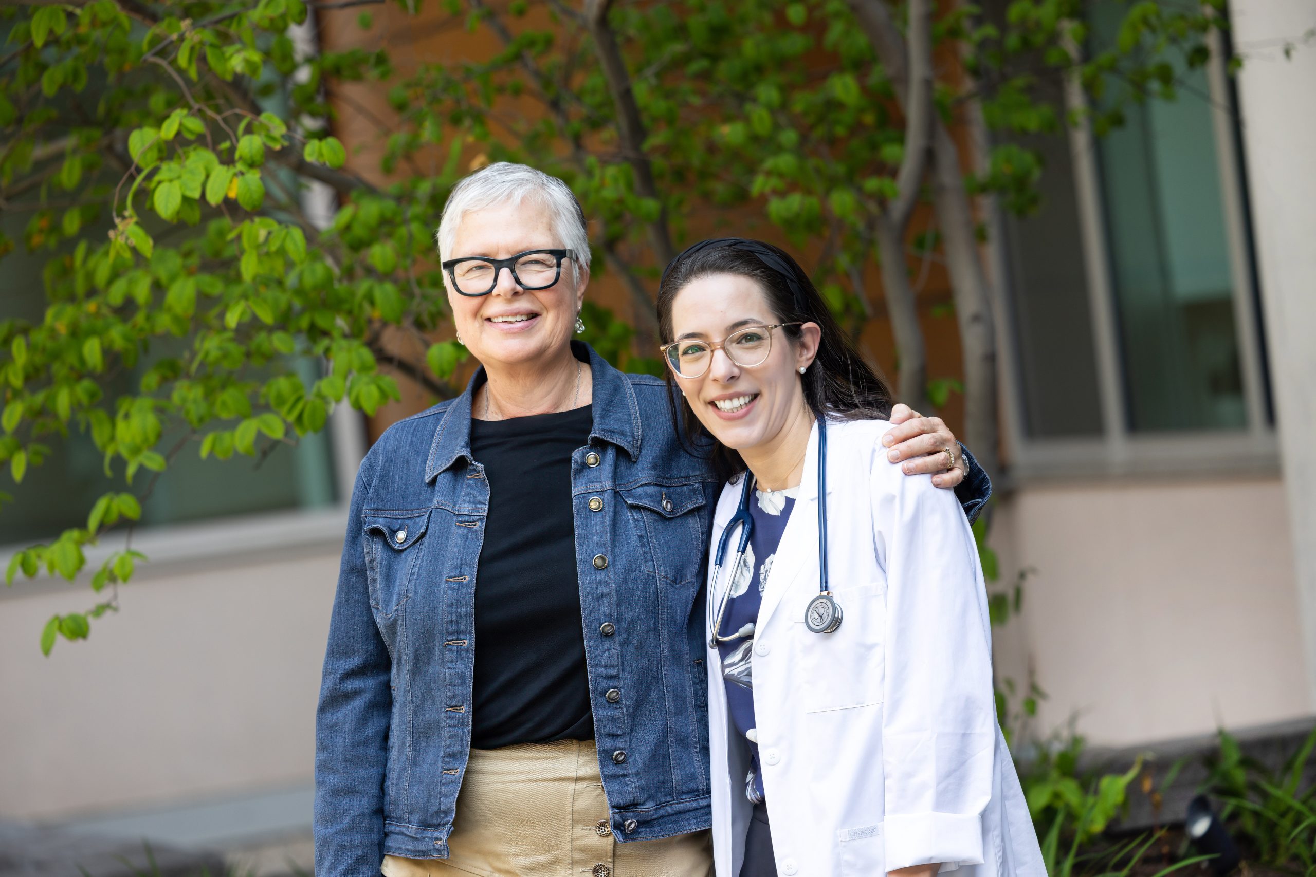 Two women stand together outdoors, smiling. One wears a denim jacket, the other a lab coat and stethoscope.