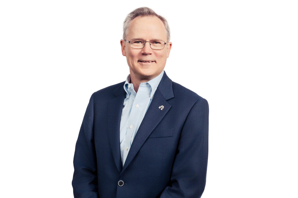 Smiling man in a navy suit, light blue shirt, standing against a white background.