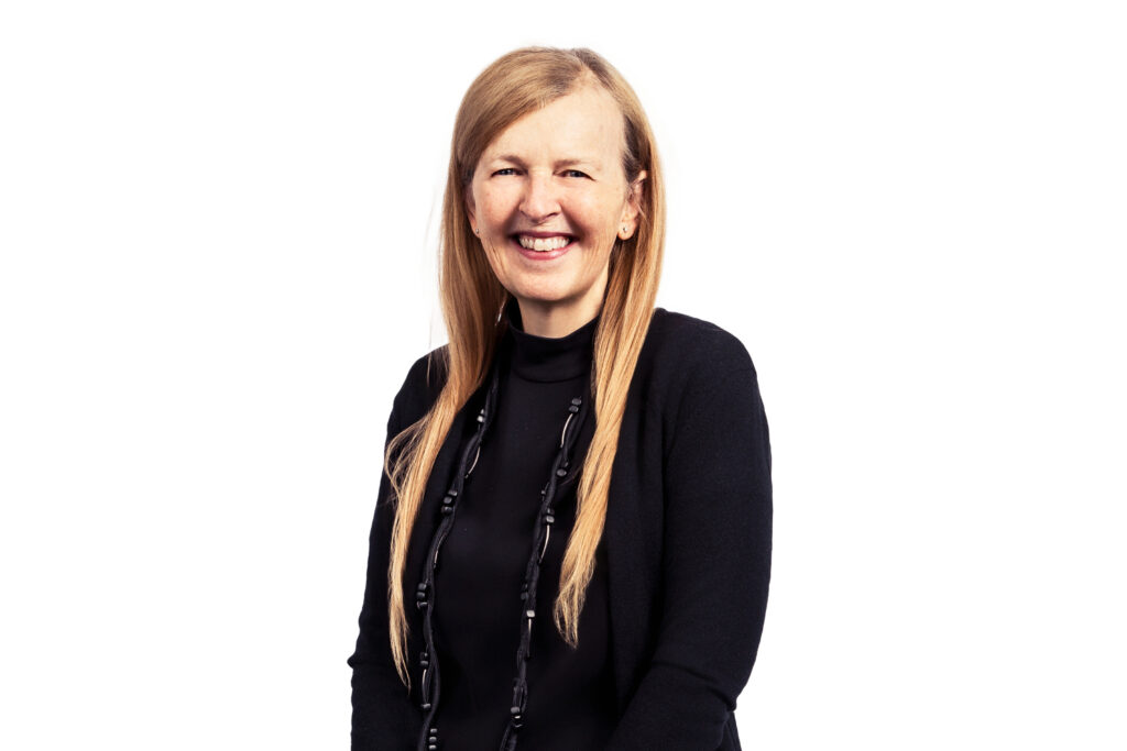 Smiling woman with long blonde hair, wearing a black top and beaded necklace, against a white backdrop.